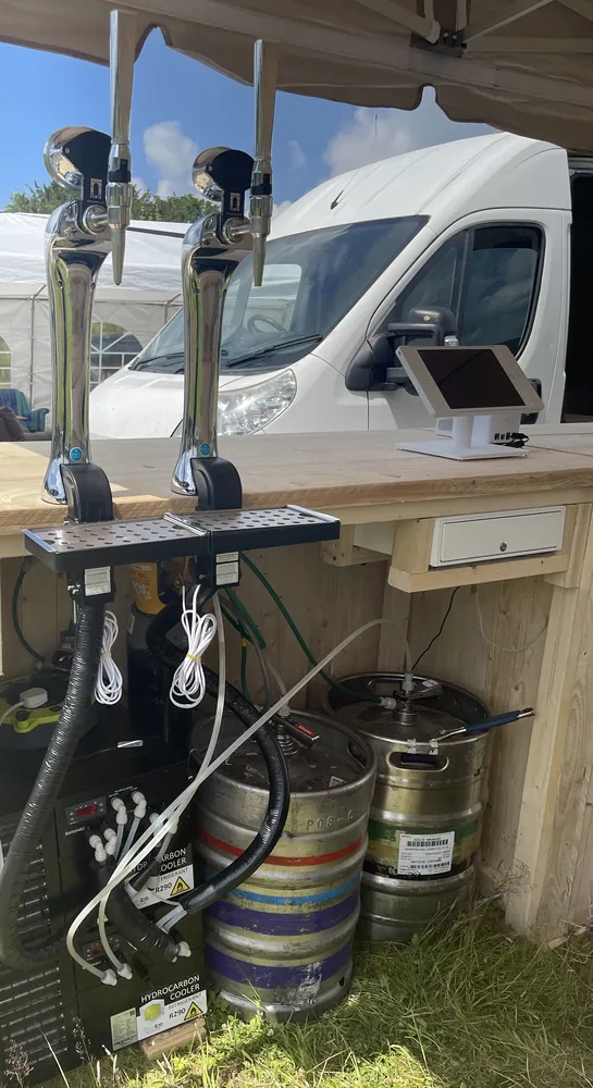 Two chrome taps above a cooler, two kegs, beer lines, drip trays and a till drawer under a field event bar